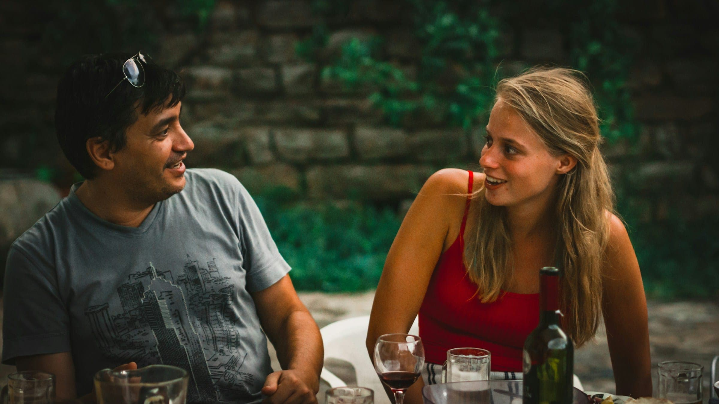 A man and a woman smiling at each other over wine at an outdoor dinner date