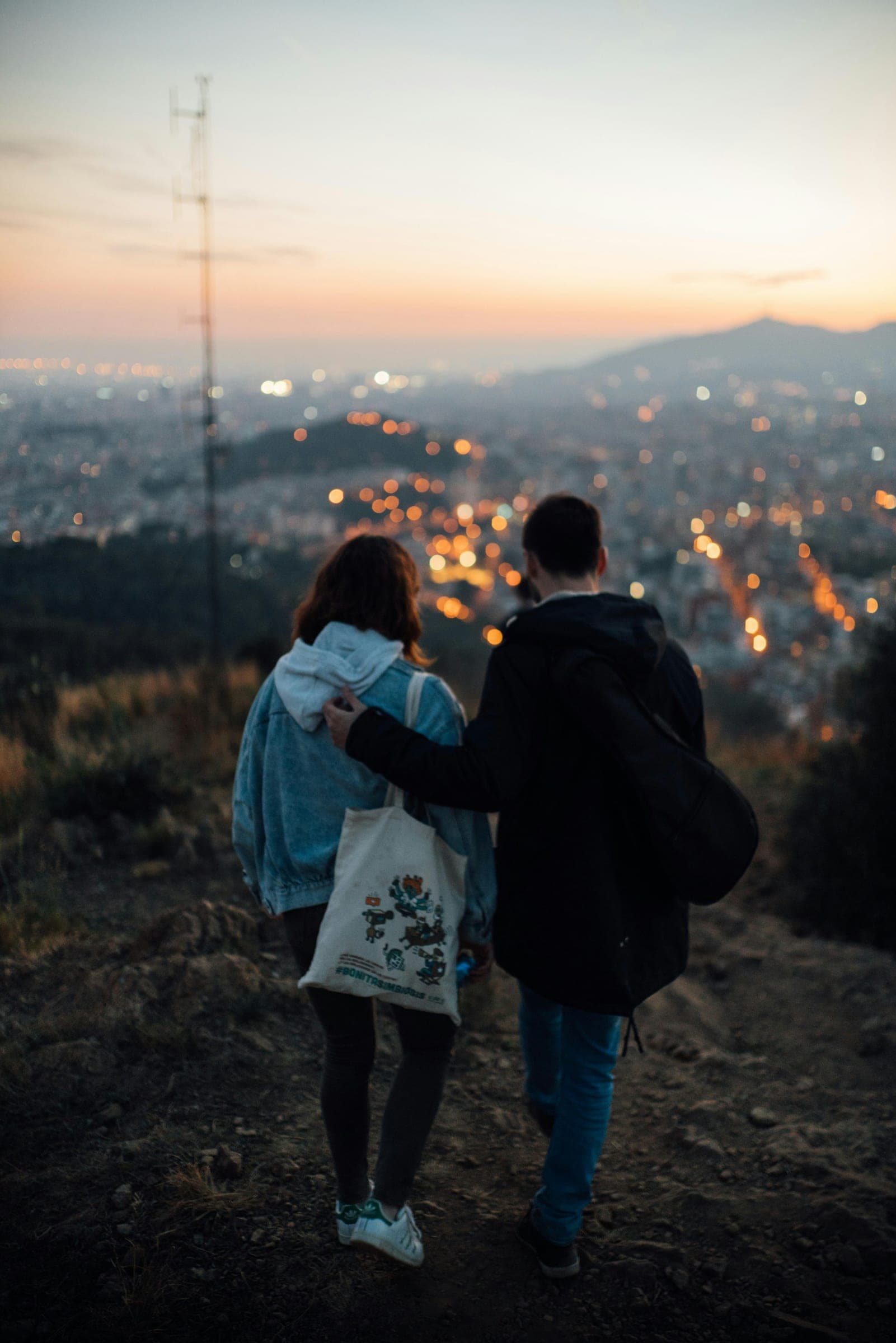 A couple walking arm in arm on a hilltop above the lights of Barcelona at dusk