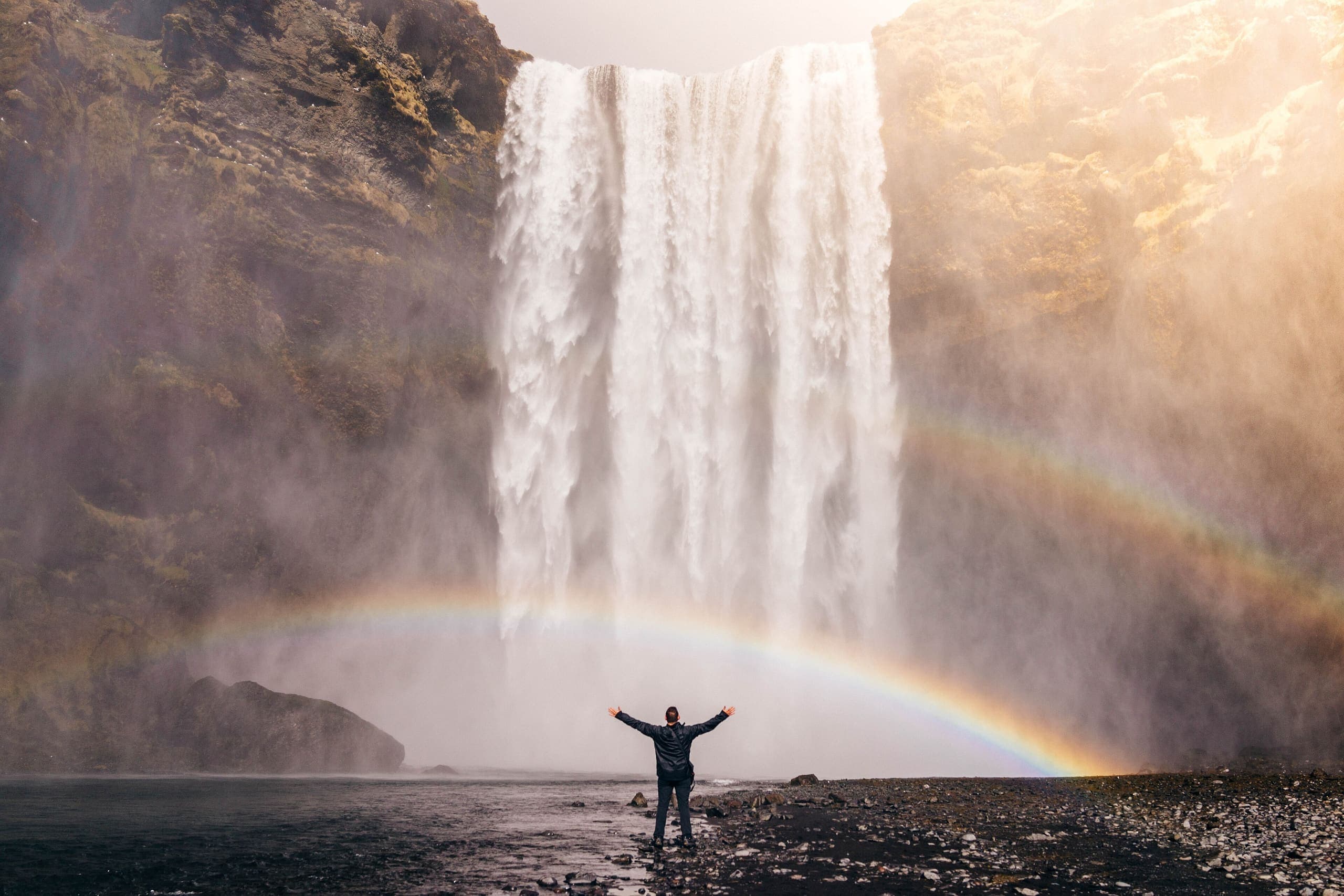 Traveler with arms raised below a large waterfall with a rainbow in Iceland