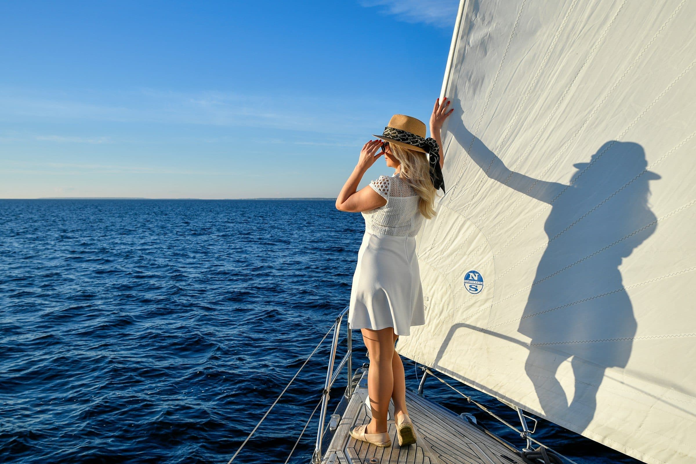 A woman in a white summer dress on the bow of a sailboat, the open sea and a clear blue sky ahead